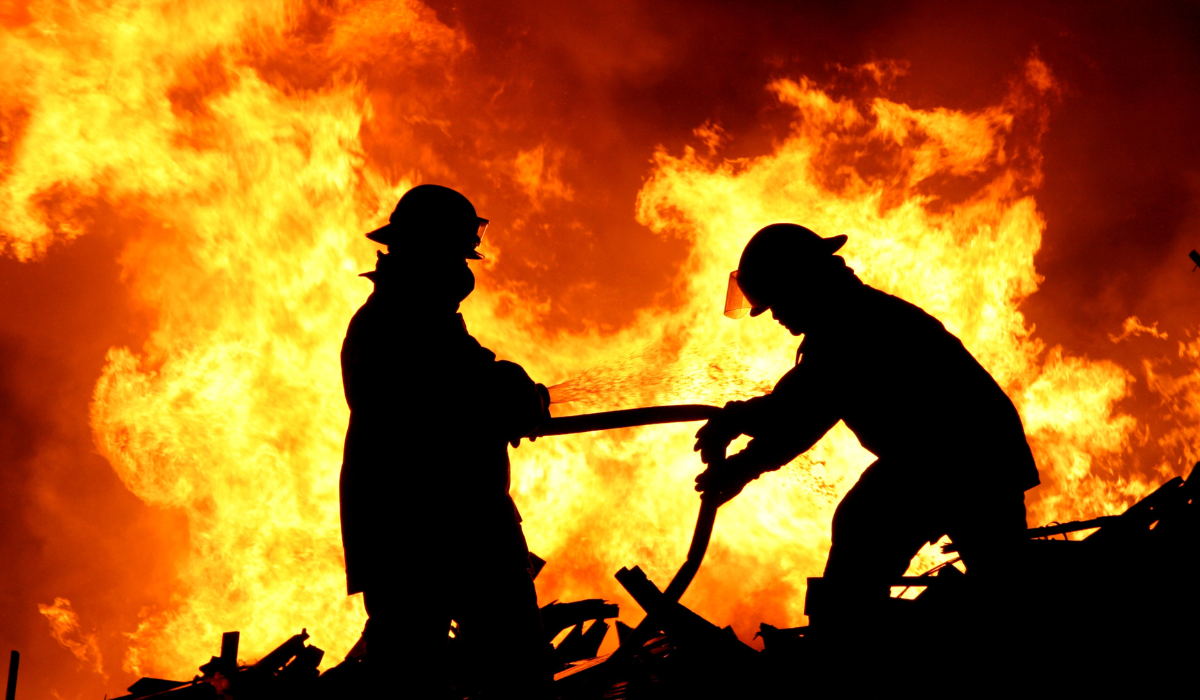 two fire fighters in full gear, silhouetted in front of a large fire 