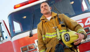A slightly angled photo of a fire fighter in uniform, standing in front of a fire truck, and holding his fire helmet. Health monitoring for emergency responders