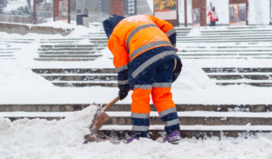 An outdoor worker in winter, shoveling snow and wearing a bright orange safety jacket