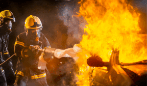 2 firefighters spraying firefighting foam onto a burning car, exposing them to PFAS